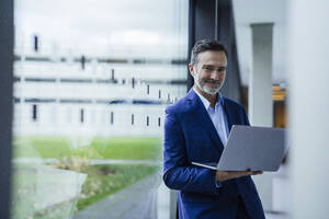 Lächelnder Geschäftsmann hält Laptop in der Nähe eines Glasfensters im Bürokorridor - JOSEF22483