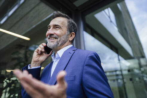 Lächelnder Geschäftsmann, der im Büro gestikuliert und mit seinem Smartphone spricht - JOSEF22488