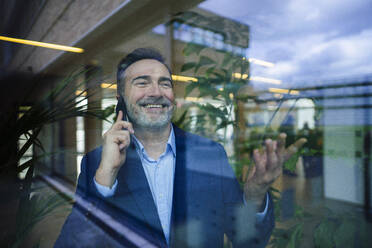 Lächelnder Geschäftsmann, der gestikuliert und mit seinem Smartphone spricht, gesehen durch Glas - JOSEF22492