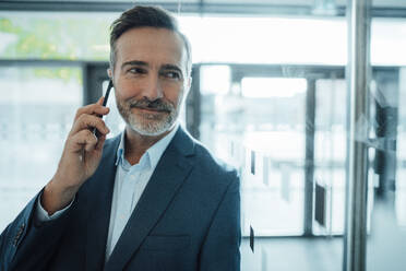 Lächelnder Geschäftsmann, der mit seinem Smartphone im Büro spricht - JOSEF22494