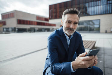 Lächelnder Geschäftsmann sitzt mit Smartphone im Büropark - JOSEF22495