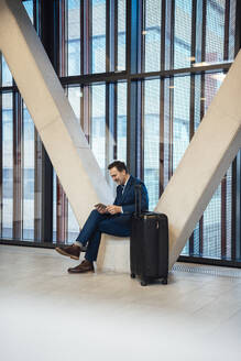 Geschäftsmann sitzt mit Koffer und benutzt Tablet-PC in der Büro-Lobby - JOSEF22504