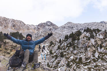 Lächelnder Mann sitzt mit ausgestreckten Armen auf einem Felsen vor den Pyrenäen in Katalonien, Spanien - PCLF00894