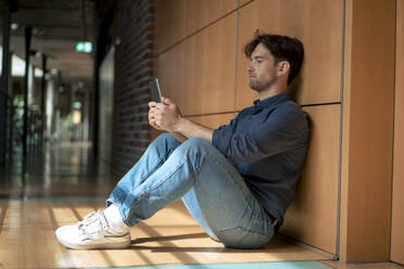 Young businessman using tablet PC sitting in corridor at office - JOSEF22526