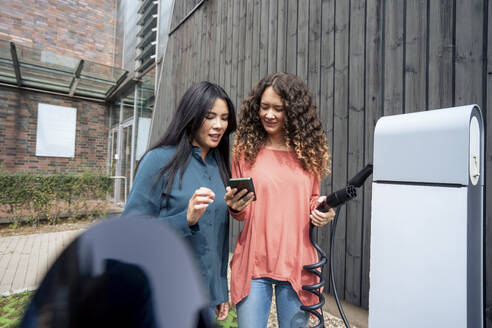 Woman holding electric plug and sharing smart phone with friend at charging station - JOSEF22545