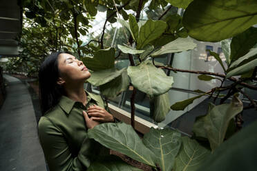 Businesswoman with hands on chest standing by plants in corridor - JOSEF22599