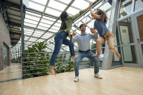 Multiracial businesswomen standing on colleague's legs and giving high-five in corridor - JOSEF22609
