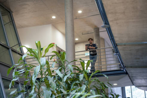 Businessman using tablet PC and leaning on railing of office corridor - JOSEF22622