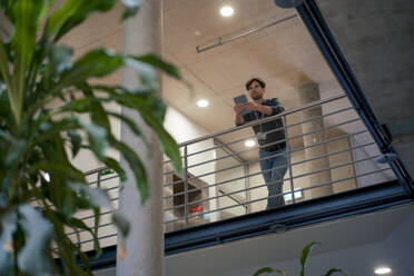 Young businessman with tablet PC leaning on railing of office corridor - JOSEF22624