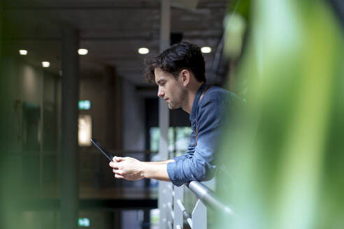 Businessman using tablet computer leaning on railing in corridor - JOSEF22628