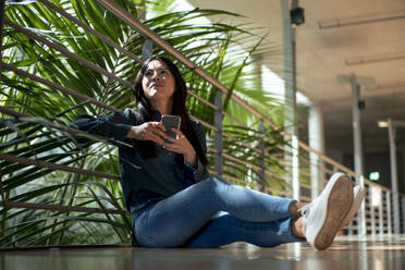 Thoughtful businesswoman with smart phone sitting in corridor - JOSEF22643