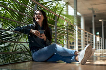 Happy businesswoman with mobile phone sitting in corridor - JOSEF22644