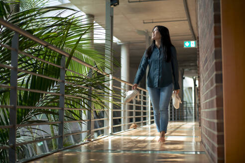 Businesswoman holding shoes and walking bare feet in corridor of office - JOSEF22655