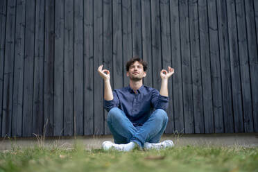 Young man meditating sitting on grass in front of wall - JOSEF22670