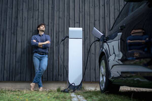 Young man with arms crossed leaning on wall by electric car - JOSEF22672