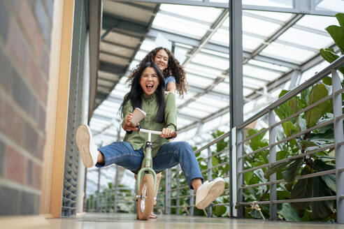 Happy businesswoman enjoys riding bicycle with colleague pushing her in corridor - JOSEF22709