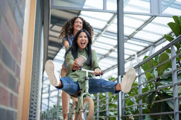 Businesswoman enjoys riding bicycle with colleague pushing her in corridor - JOSEF22710