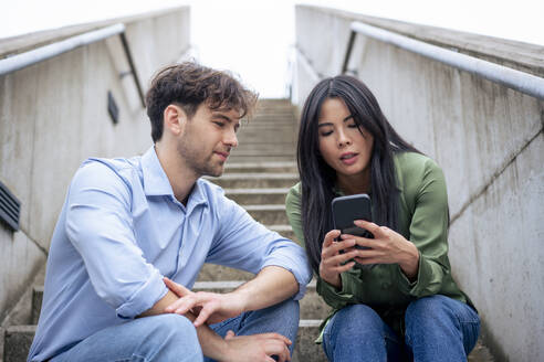 Woman sharing mobile phone with friend sitting on staircase - JOSEF22728