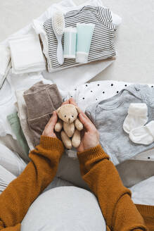 Pregnant woman holding stuffed teddy bear near clothes at home - NDEF01497
