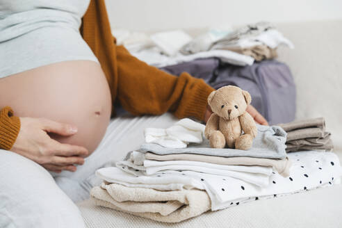 Pregnant woman sitting with stuffed teddy bear and packing clothes at home - NDEF01498