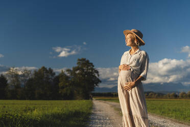Nachdenkliche schwangere Frau steht auf der Straße in der Nähe von Feld bei Sonnenuntergang - NDEF01508