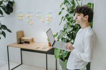 Thoughtful businessman standing with laptop in office - YTF01542