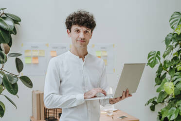 Smiling businessman holding laptop near plants in front of wall - YTF01547