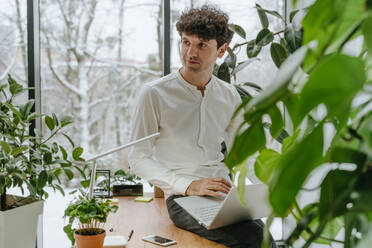 Contemplative businessman sitting with laptop on desk in office - YTF01554