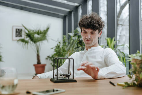 Smiling businessman playing with newton's cradle in office - YTF01572