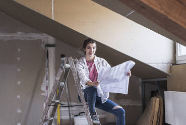 Lächelnde Frau auf Leiter sitzend mit Bauplan zu Hause - UUF30879