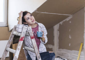 Contemplative woman holding coffee mug and leaning on step ladder at home - UUF30882