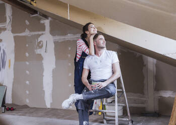 Smiling man sitting on ladder with woman standing at home - UUF30897