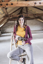 Smiling woman sitting on ladder with electric drill at home - UUF30904