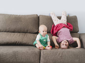 Verspieltes Mädchen macht Kopfstand in der Nähe des kleinen Bruders auf dem Sofa zu Hause - NDEF01525