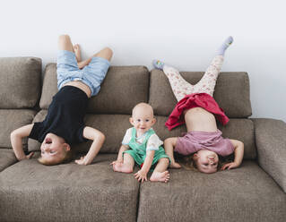 Happy children doing headstand near baby brother on sofa at home - NDEF01526