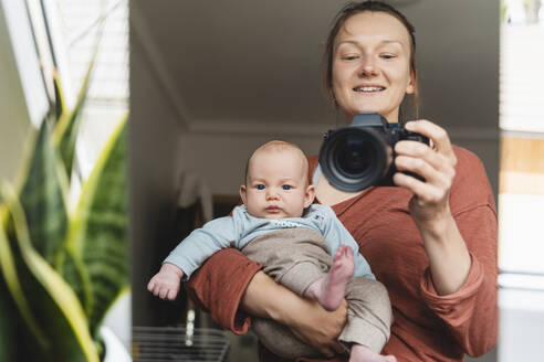 Spiegelung einer lächelnden Mutter, die ihren Sohn trägt und durch die Kamera fotografiert - NDEF01531