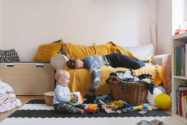 Erschöpfte Mutter schläft auf dem Sofa, während ihr kleiner Junge zu Hause spielt - NDEF01543