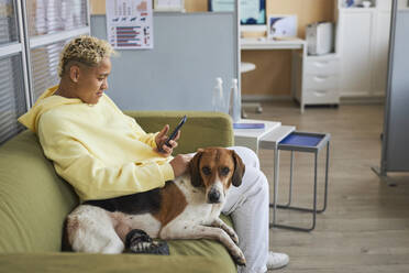 Frau, die ein Mobiltelefon benutzt und mit einem behinderten Hund auf dem Sofa in einer Tierklinik sitzt - KPEF00523