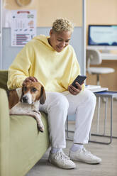 Lächelnde Frau, die ein Smartphone benutzt und einen Hund auf dem Sofa in einer Tierklinik streichelt - KPEF00525