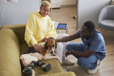 Glückliche Frau mit Tierarzt streichelt Hund auf Sofa in Klinik - KPEF00526