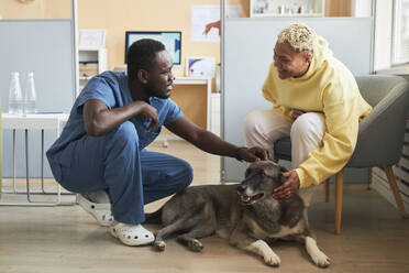 Glückliche Frau im Gespräch mit einem Tierarzt, der einen Hund in der Klinik streichelt - KPEF00531