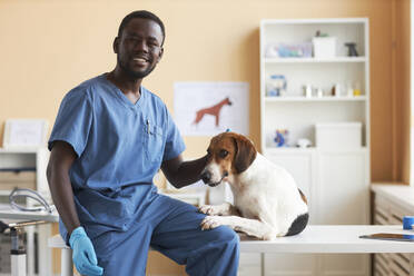 Lächelnde junge Tierärztin sitzt mit Beagle-Hund auf dem Tisch in der Klinik - KPEF00552