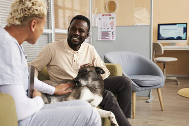 Happy man sitting with dog and talking to veterinarian in clinic - KPEF00560