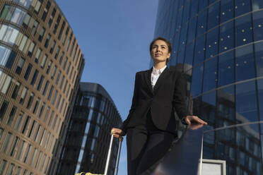 Smiling businesswoman with suitcase standing near buildings in city under sky - VPIF09205