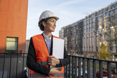 Smiling engineer holding file folder and laptop near railing in city - VPIF09226