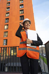 Happy engineer standing with file folder and laptop in city - VPIF09228