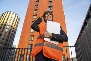 Ein lächelnder Ingenieur steht mit Laptop und Aktenordner vor einem Gebäude - VPIF09229
