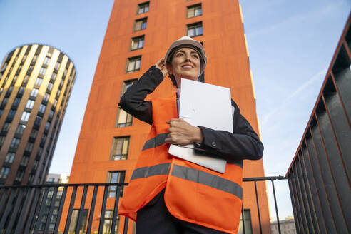 Smiling engineer standing with laptop and file folder in front of building - VPIF09229