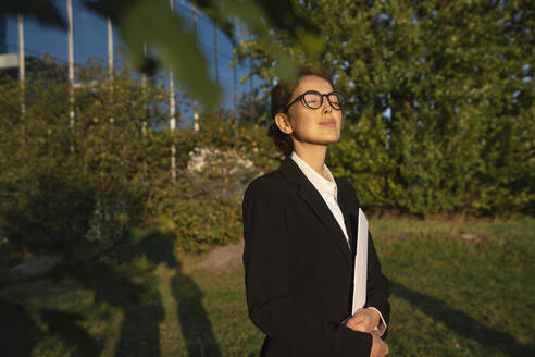 Businesswoman standing with file folder and eyes closed in office park - VPIF09238