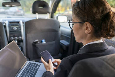 Businesswoman using mobile phone near laptop in car - VPIF09253